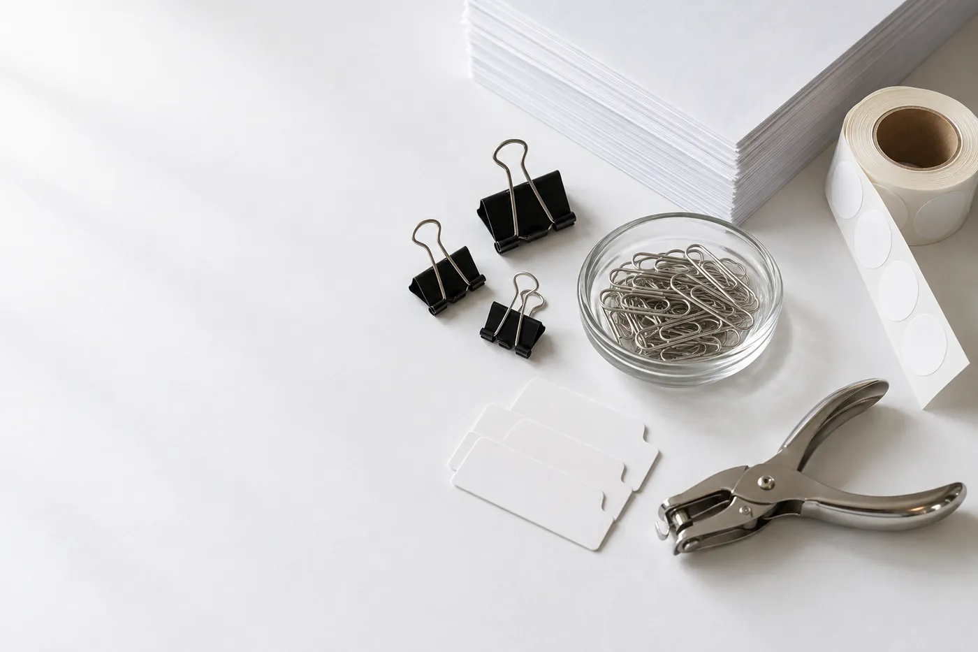 Illustration: Foldback-Klammern, Büroklammern in einer Glasschale, eine Rolle runder Etiketten, Registerschildchen und ein Locher neben einem Stapel Druckerpapier
